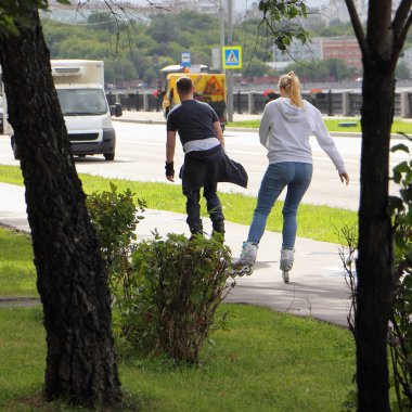 Rus genç aile patencileri, sıradan giysiler içinde yaz günü manzarasında boş kaldırım kenarında paten kayarak geziyorlar. Moskova 'da şehir sporları.