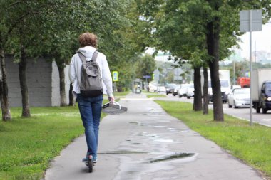 Avrupa 'da yaz günü sırt çantalı, elektrikli scooterlı bir adam boş asfalt sokağın sol tarafında, arka manzaralı.