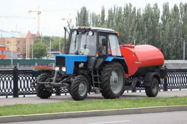 Mavi Belarus traktör sulama makinesi turuncu fıçı ile asfalt dolgu kaldırımı, Moskova 'da sokak yıkama güneşli bir yaz gününde belediye hizmetleri tarafından şehir iyileştirme