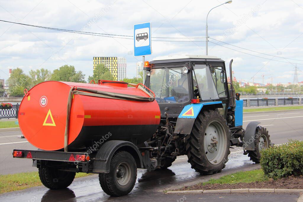 Blue Belarus tractor watering machine street cleaner with orange barrel pours asphalted  sidewalk, street washing in Moscow, city improvement by municipal services on a sunny summer day