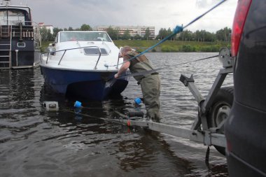 Beyaz mavi kabin motorlu teknesi kalkış yapıyor, beyaz bir adam yaz günü kayıktaki siyah araba römorkuna bir tekne yerleştiriyor.