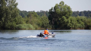 Turuncu can yelekli bir adam hızlı bir şekilde şişirilebilir motorlu teknede, yeşil ağaçların üzerinde, arka planda, Sunnny 'nin yaz günü sularında yüzer.
