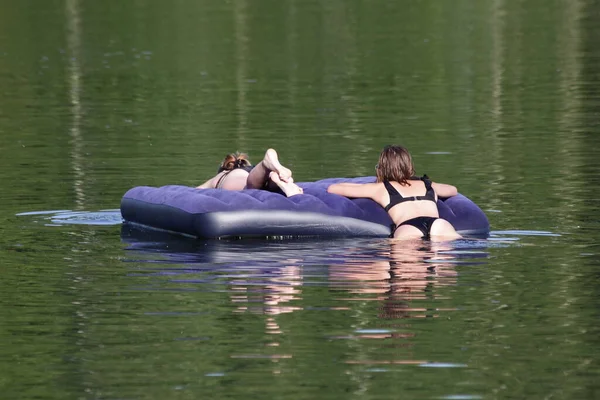 二人の白人の女の子は池の青いインフレータブル空気のベッドで泳いでいます 夏の日に水の上の蒸気の安全性 — ストック写真