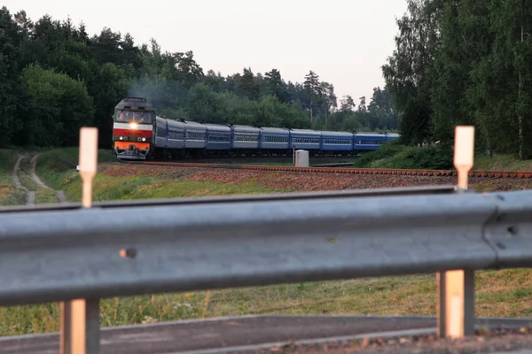 Old approaching passenger train, a view from a car against the background of a railway crossing barrier and trees