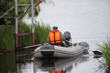 Turuncu can yeleği giymiş bir balıkçı, sakin suda motoru kırık gri şişirilebilir bir motorla kürek çeker. Yaz sabahı balık tuttuktan sonra balıkçı üssü girişine döner.