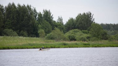 Rus kırsal nehir manzarası, sakin nehir suyunda yüzen eski motorlu tekne, yeşil çimenli sahil yaz günü orman geçmişi, doğada dinlenme.