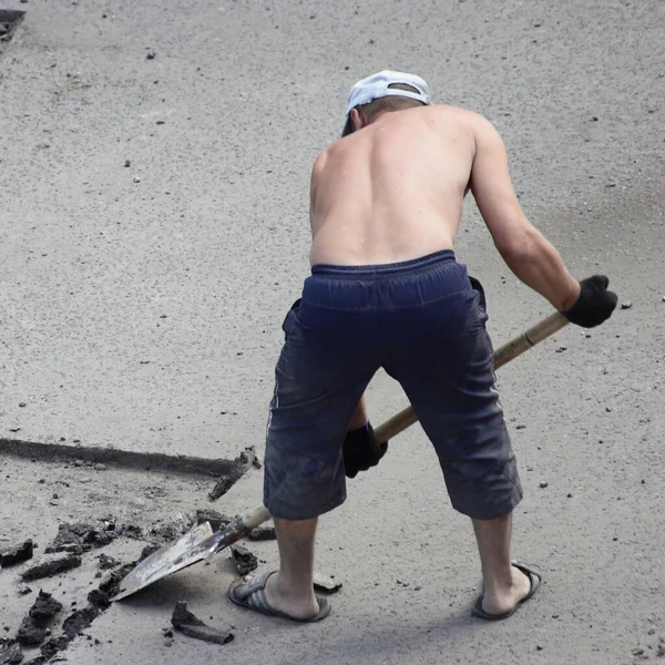防護服のない一人の白人道路労働者は 晴れた夏の日にシャベルで古いアスファルトを取り除き 道路の便利な修理重い仕事を閉じます — ストック写真