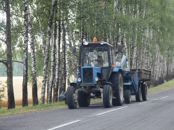 夏の日に村のバーチのトレーソンフィールドの背景の間の田舎のアスファルトの道路上のトレーラードライブと車輪付きトラクター 東ヨーロッパでの収穫 — ストック写真