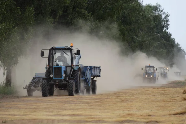 夏の日に東ヨーロッパでのリネンフィールドエッジ道路 リナム収穫上のほこりの雲の中に高速ドライブの収穫トレーラーを持つ多くのトラクター — ストック写真