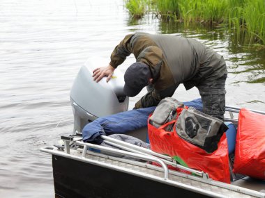 Boater man checks the hood of a 50 hp four stroke outboard motor on transom of boat near grass, repair and maintenance boat engine clipart
