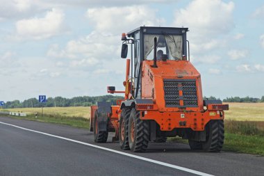 Büyük turuncu tekerlekli buldozer traktörü tarlada asfalt yol kenarında, mavi gökyüzü ve ormanın arka planında yaz gününde, Batı Avrupa çiftlik arazisinde