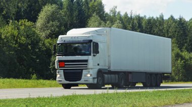 Bir Avrupa beyaz tırı ön tarafta Sunny yaz gününde banliyö otobanında yaklaşan trafiğe bakıyor yeşil orman arka planı, kargo taşımacılığı lojistik trafik manzarası