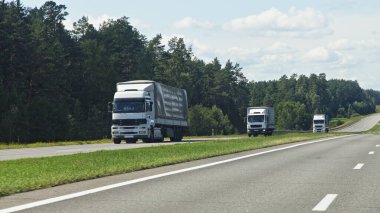 Avrupai kamyonlar, yeşil orman ve mavi bulutlu gökyüzü arka planı, trafik manzarası üzerinde Sunny yaz gününde banliyö otobanında yaklaşan trafiğe ön taraftan bakıyorlar.