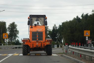 Büyük, 3 eksenli turuncu tekerlekli buldozer traktörü, yaya geçidinde yaz günü demiryolu kavşağından önce, endüstriyel makine trafiği yolda.