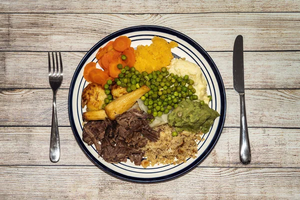 Sunday dinner of slow cooked shredded pulled beef on a plate with a knife and fork and plenty of vegetables such as peas, mushy peas, parsnips, roast potatoes and carrots, stock photo image