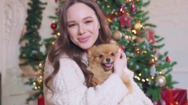 Beyaz kazaklı genç bir esmer, uzun saçlı, elinde kırmızı bir Spitz köpeği var. Arka planda, bir Noel ağacının yanında. Gülümsüyor, köpeğe sarılıyor.