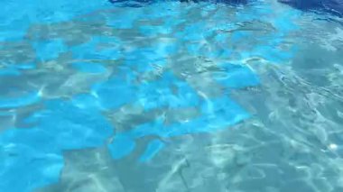 Açık havuz alanında. Güneş ışığı berrak suda parlıyor ve havuz zemininde çeşitli tonlarda mavi fayanslar var..