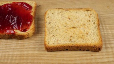 Fıstık ezmesi bıçakla ahşap bir tahtanın üzerinde çoklu tahıllı tost ekmeğine sürülür ve çift fıstık ezmeli ve kırmızı üzümlü reçelli sandviç yapılır..