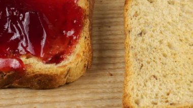 Fıstık ezmesi bıçakla ahşap bir tahtanın üzerinde çoklu tahıllı tost ekmeğine sürülür ve çift fıstık ezmeli ve kırmızı üzümlü reçelli sandviç yapılır..