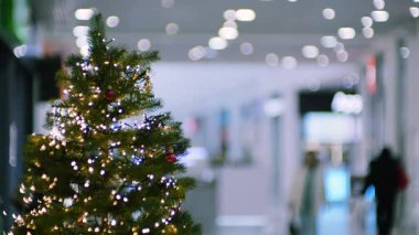 Bir Noel ağacı bir alışveriş merkezinde ışık ve süslemelerle süslenir. Bulanık bir insan figürü kalabalık, parlak koridor alışveriş merkezinde yürüyor..