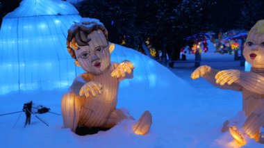 LVIV, UKRAINE - Ocak 06, 2026: Aydınlatılmış buz heykelleri büyülü bir kış harikalar diyarı yaratır. Karda oturan parlak bebek figürleri. Parkta çekim..