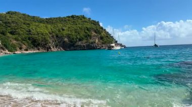Turkuaz mavi suları olan kumlu bir sahil, Saint Martin Adası 'ndaki yemyeşil kayalıklara ve demirlemiş yelkenlilere bakar..