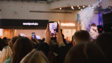 Genç hayranlar bir alışveriş merkezinin avlusunda akıllı telefonlardan konser çekiyorlar. Büyük bir gençlik kalabalığı sahnenin yanında toplandı. Enerji dolu izleyiciler halka açık bir etkinlikte konser veriyor..