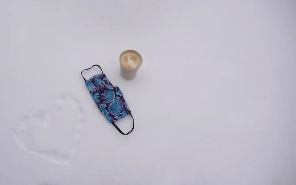 COVID19 настоящий. Медицинская маска и чашка кофе на белоснежном снегу. Блокировка и отдых от карантина. — стоковое фото