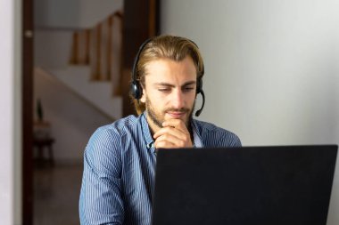 Evde kulaklıkla çalışan sarışın bir erkek dizüstü bilgisayarda kablosuz internet kullanıyor, erkek çağrı merkezi temsilcisi ya da tele pazarlamacı.