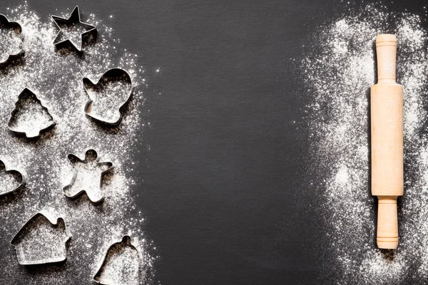 Christmas cookies molds, rolling pin and flour on black background with copy space.