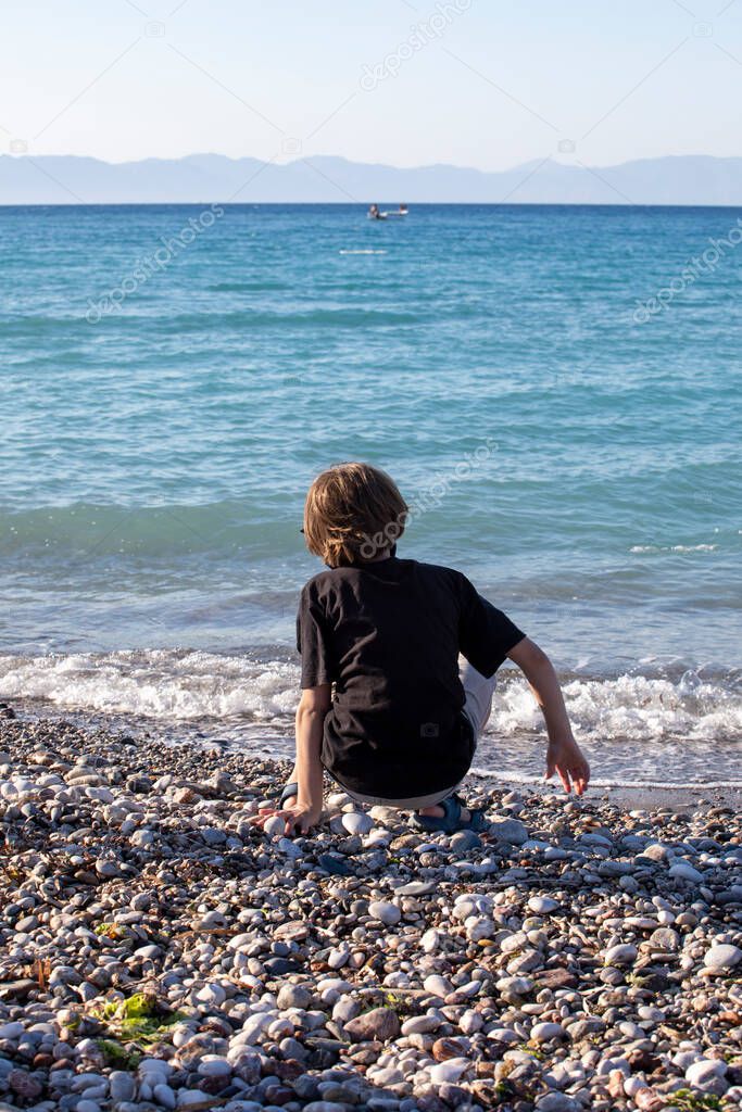 happy boy with his back sits on the seashore and throw stones family vacation, greec