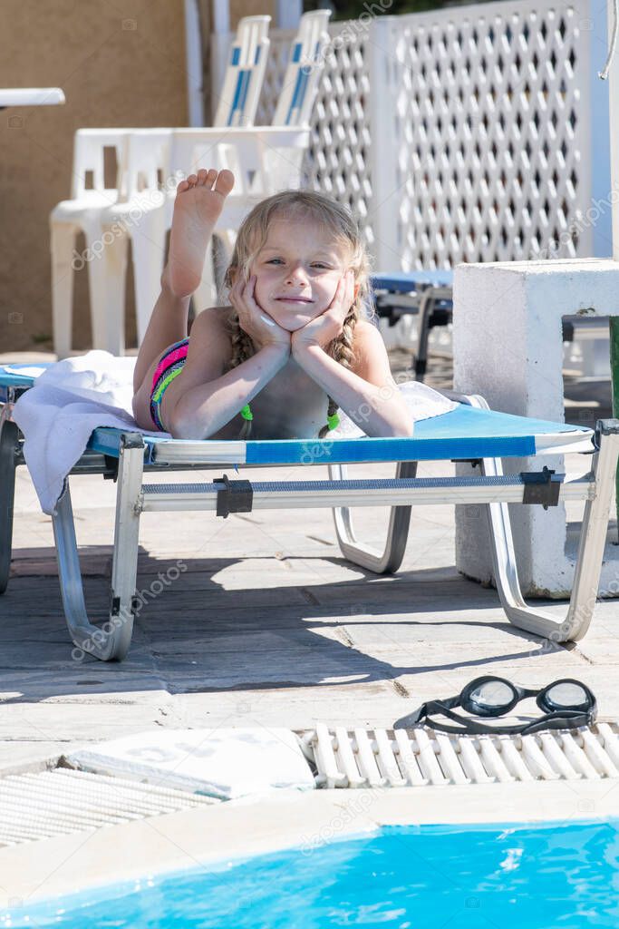 little happy girl lies on a sun lounger in the shade and sunbathes by the pool, family vacation Greece