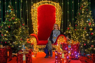 Noel ağaçları ve hediye kutuları arasında kırmızı bir tahtta oturuyor.