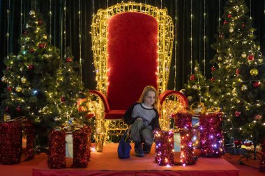 Noel ağaçları ve hediye kutuları arasında kırmızı bir tahtta oturuyor. Dilek tutuyor.