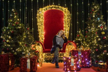 Noel ağaçları ve hediye kutuları arasında kırmızı bir tahtta oturuyor. Dilek tutuyor.