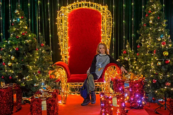 Noel ağaçları ve hediye kutuları arasında kırmızı bir tahtta oturuyor.
