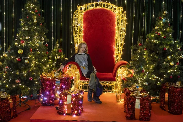 Noel ağaçları ve hediye kutuları arasında kırmızı bir tahtta oturuyor.