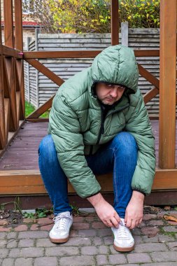 Bir adam terasta ceketinin içinde oturur ve dışarıya ayakkabı bağı bağlar..