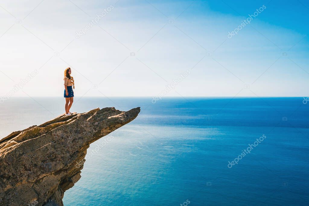 A young woman stands on a picturesque rock ledge above the sea against the sky. The concept of travel and freedom.