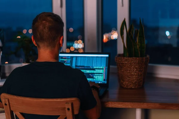 A young man programmer coding on a laptop in the dark with a view of the lights of the night city, color lighting in the room, home decor