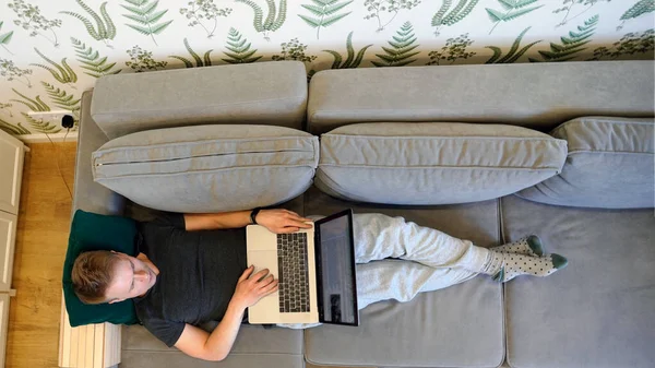 若い男が膝の上にノートパソコンを持って家のソファに横になっている 家庭から働くという概念 写真を挿入するための緑色の画面 上からの写真 — ストック写真