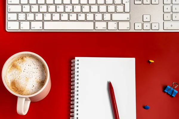 Top view of the office desk. A workspace with an empty notebook, keyboard, stationery, and coffee cup on a red background.
