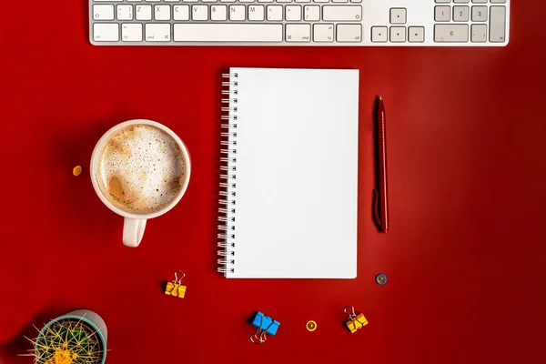 Top view of the office desk. A workspace with an empty notebook, keyboard, stationery, and coffee cup on a red background.