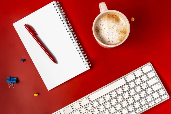 Top view of the office desk. A workspace with an empty notebook, keyboard, stationery, and coffee cup on a red background.