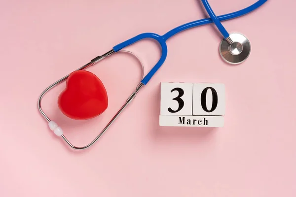 The concept of celebrating Doctor\'s Day with a stethoscope and a heart on a pink background. Wooden calendar with the date March 30