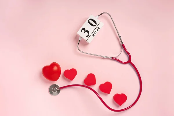 The concept of celebrating Doctor\'s Day with a stethoscope and a heart on a pink background. Wooden calendar with the date March 30