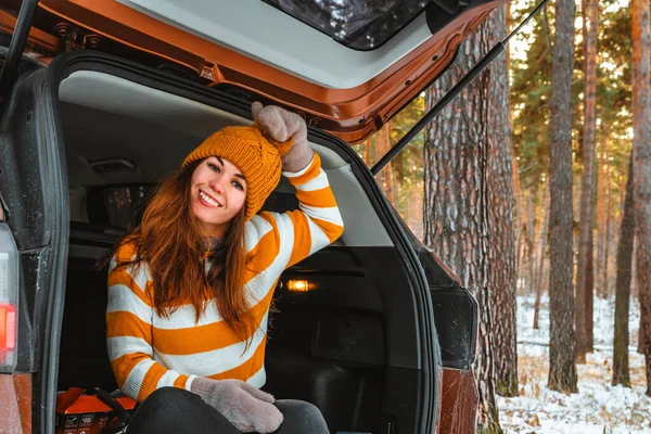 A smiling woman in a sweater and hat sits in the open trunk of a car against the background of a snowy landscape in a winter pine forest, road trip