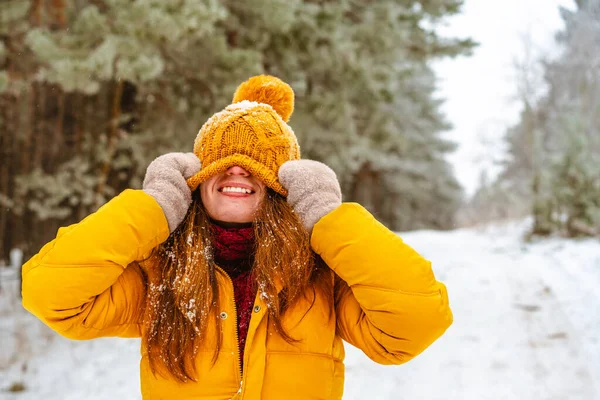 Stylish happy girl with yellow clothes is fooling around and pulling a winter hat over her face on winter Christmas vacation in the forest