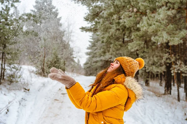 黄色のジャケットと帽子の美しい若い女性は雪の冬の森の中で雪を投げる — ストック写真