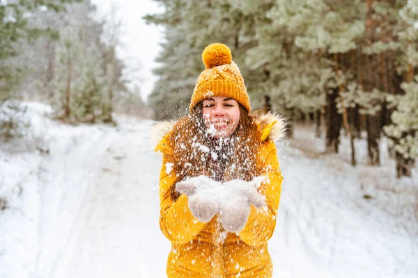 黄色のジャケットと帽子の美しい若い女性は雪の冬の森の中で雪を投げる — ストック写真